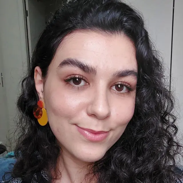 Foto de perfil da psicóloga Amanda Bonafini, com cabelo castanho escuro, ondulado e comprido, brincos médios nas cores amarela e alaranjada. Ela está sorrindo levemente.