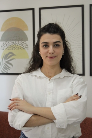 Foto de perfil da psicóloga Amanda Bonafini, com cabelo castanho escuro, ondulado e comprido, brincos médios nas cores amarela e alaranjada. Ela está sorrindo levemente.